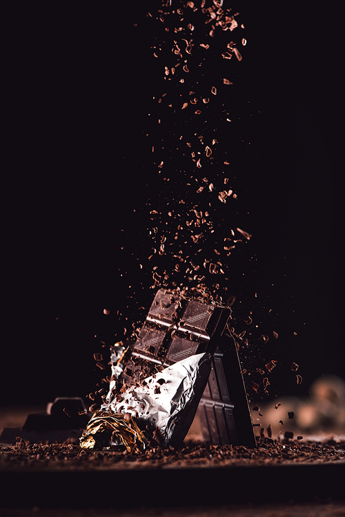 close up image of grated chocolate falling on two chocolate bars on wooden table on black background