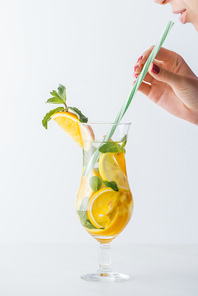 partial view of woman holding straw and summer cold cocktail with mint, lemon and orange pieces isolated on white