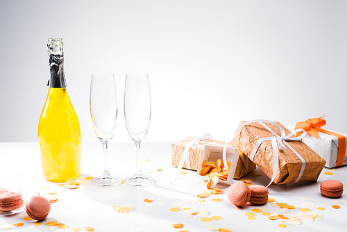 close up view of bottle of yellow champagne, empty glasses, macarons and arranged gifts on grey backdrop