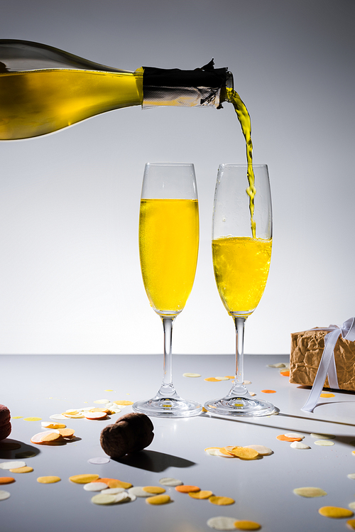 close up view of pouring yellow champagne into glasses process and wrapped gift on grey backdrop