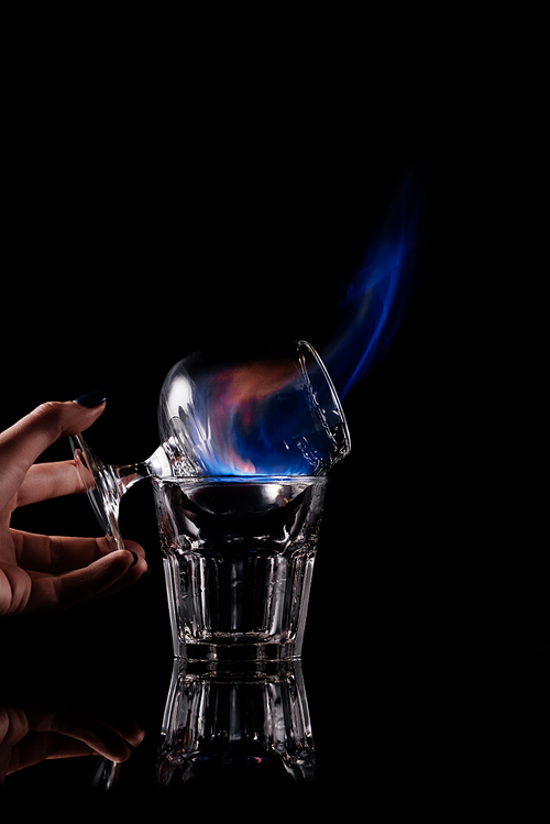 partial view of woman holding glass with burning sambuca alcohol drink on black background