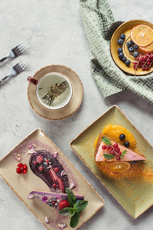 flat lay with sweet carrot cake with berry filling, blueberry cake served with mint leaves and violet petals, cup of herbal tea and cutlery on grey tabletop