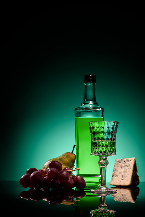 close-up shot of absinthe with pear, grapes and cheese on mirror surface on dark blue background