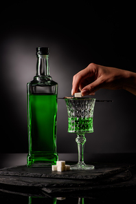 cropped shot of woman putting sugar cube on absinthe spoon on glass on dark background