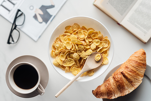 top view of bowl of dry cereal breakfast with cup of coffee and croissant on white surface with newspaper and book