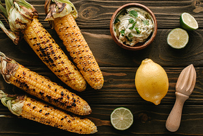 top view of grilled corn, lime slices, lemon, wooden squeezer and cream with parsley on wooden table
