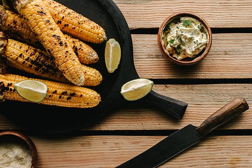 top view of grilled corn, lime slices, knife and butter with parsley on wooden table