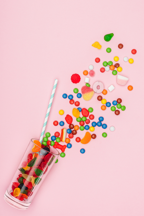 top view of scattered jelly and dragee candies, glass and plastic straw isolated on pink