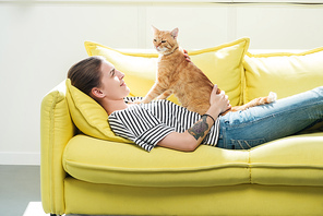 beautiful girl lying on yellow sofa and palming cute red cat in living room