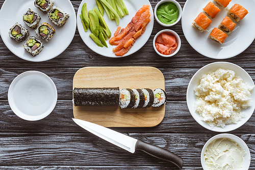 top view of delicious sliced sushi roll, knife and ingredients on wooden table