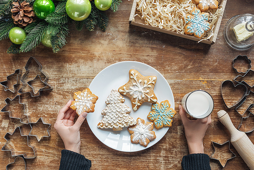 partial view of woman holding glass of milk and cookie on wooden tabletop with decorative christmas wreath and cardboard box