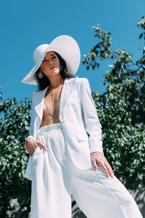 low angle view of attractive woman in white suit and hat posing outside