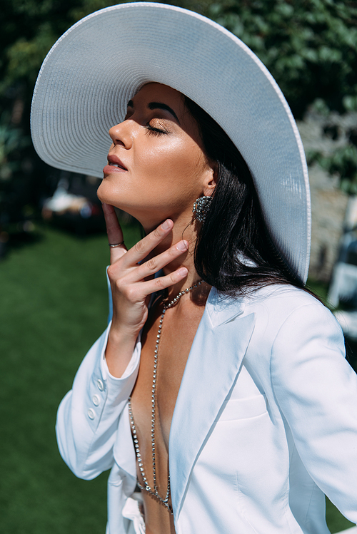 attractive woman in white suit and hat posing with closed eyes outside