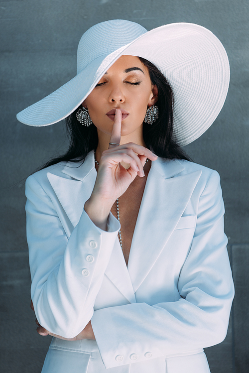 attractive woman in white suit and hat posing and showing shh gesture outside