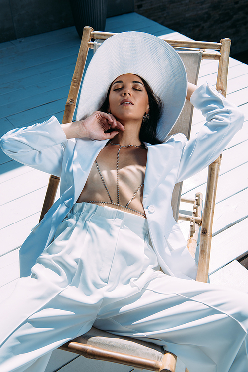 attractive woman in white suit and hat sitting om deck chair with closed eyes outside
