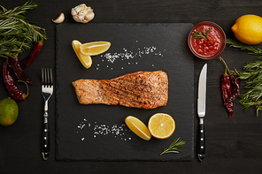 top view of grilled salmon steak with pieces of lemon, arranged ingredients around and cutlery on black surface
