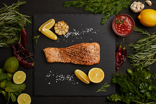 top view of grilled salmon steak with pieces of lemon and arranged ingredients around on black surface