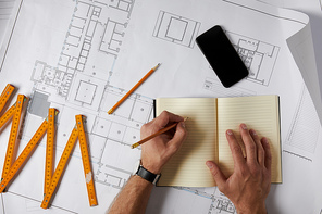 cropped image of architect writing in blank textbook on blueprint with pencil, smartphone and collapsible meter