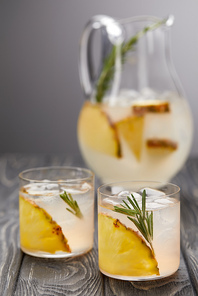 jug of lemonade with pineapple and rosemary, two glasses of lemonade with pineapple pieces, ice cubes and rosemary on grey wooden tabletop