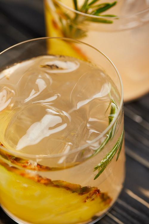selective focus of two glasses of lemonade with pineapple pieces, ice cubes and rosemary on grey wooden tabletop