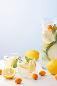 jug and two glasses of lemonade with mint leaves, ice cubes and lemon slices surrounded by kumquats and lemons on blue background