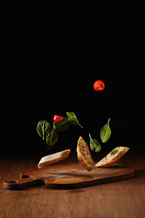 Bread pieces and salad leaves with tomatoes flying above wooden table surface
