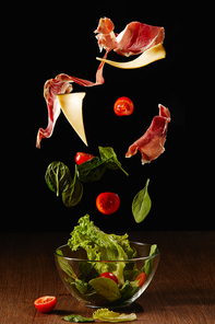 Ingredients for salad of vegetables and jamon falling in bowl above wooden table