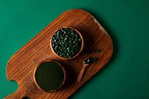 top view of cutting board, wooden spoon,  bowls with spirulina powder and spirulina in pills on green table