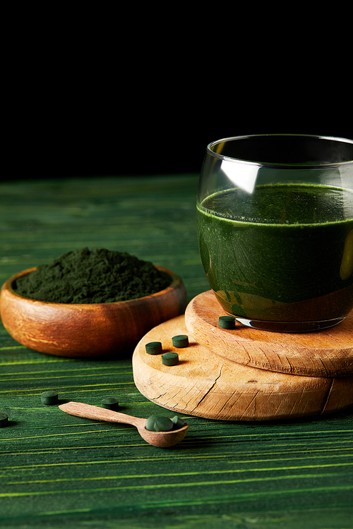 wooden slices with fresh spirulina drink in glass, wooden spoon and spirulina powder in wooden bowl on black background on green wooden table