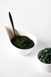 close up view of spoon, spirulina powder and spirulina pills in bowls on grey background