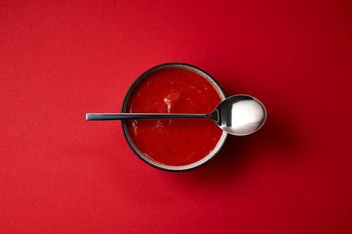top view of delicious red tomato soup in plate and spoon on red table