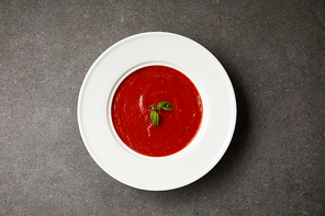 top view of red tomato soup with basil in white plate on grey table