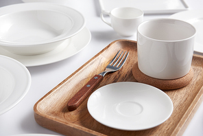selective focus of wooden tray, fork with various plates, bowl and cup on white table