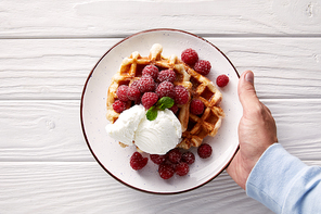 cropped shot of man holding plate of belgian waffles with raspberries and ice cream over white wooden table
