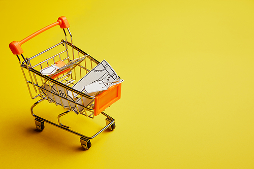 close up view of little shopping cart with clothes made of paper on yellow background