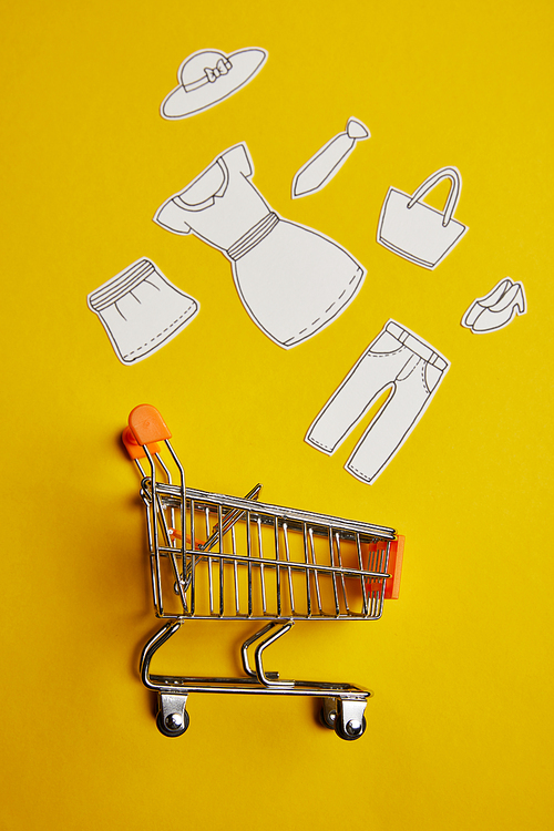 top view of little shopping trolley with paper clothes on yellow backdrop