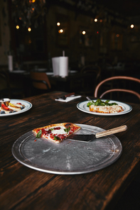 slice of pizza with server on metal tray and wooden table with plates of lasagne and syrniki
