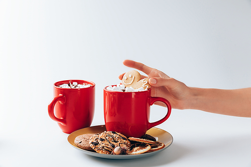 cropped view of hand puting gingerbread man into cup of hot cacao with marshmallows, on white