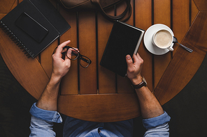 top view of man sitting at wooden table with eyeglasses, notepads, smartphone, cup of coffee and leather bag