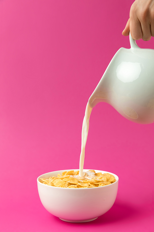 partial view of person pouring fresh healthy milk from jug into bowl with corn flakes isolated on pink