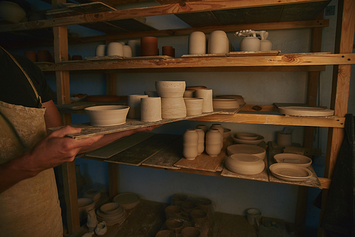 cropped image of potter in apron putting ceramic bowls and dishes on shelves at workshop
