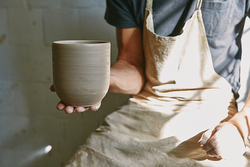 cropped image of professional potter in apron holding clay pot at workshop