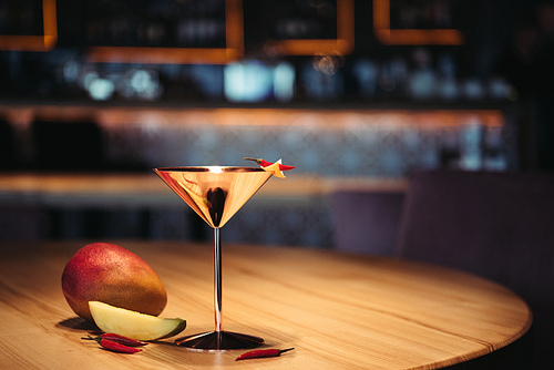 selective focus of alcoholic cocktail in metal glass decorated with chili pepper, nacho chip and mango on wooden table