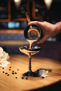 bartender pouring cocktail from shaker through sieve in metal glass on wooden table