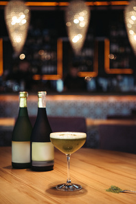 selective focus of delicious cocktail with foam on wooden table with bottles of champagne and copy space on background