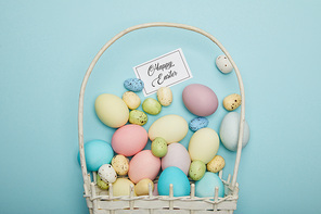 Top view of painted easter chicken and quail eggs in straw basket, card with happy easter lettering