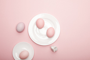 Top view of toy bunny, painted easter eggs on saucer and plate on pink background
