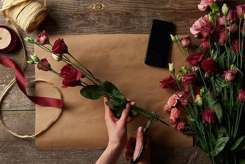 cropped shot of woman cutting flowers above wooden table with craft paper and smartphone