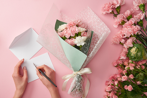 cropped shot of woman writing on greeting card and beautiful pink flower bouquet isolated on pink