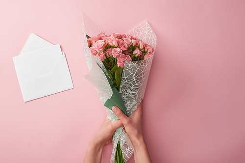 cropped shot of woman holding bouquet of beautiful roses and white envelope isolated on pink
